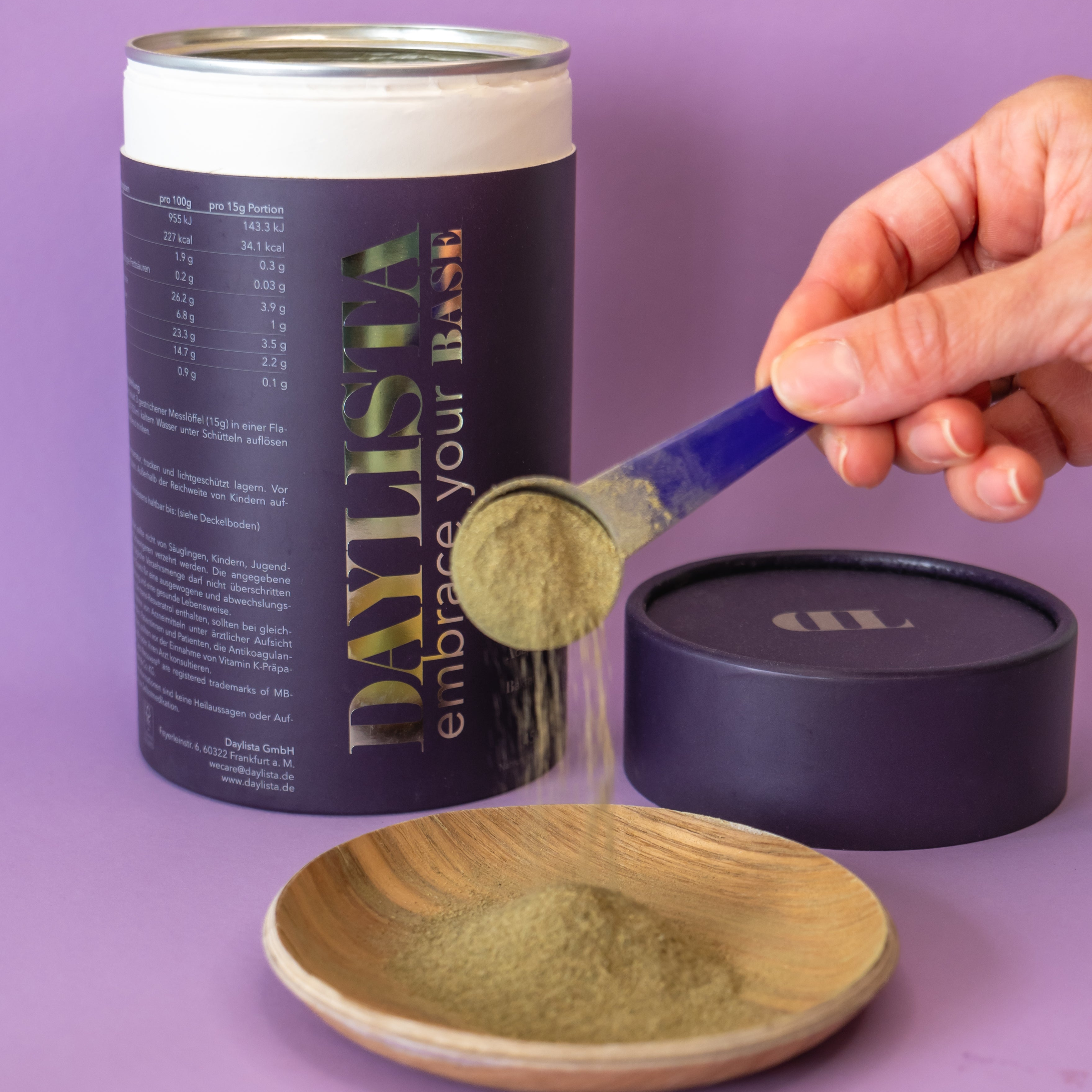 Hand hält einen Messlöffel mit beige-grünen Daylista-Pulver, das in eine Holzschale geschüttet wird; daneben steht eine geöffnete dunkelviolette Daylista-Dose vor lila Hintergrund.