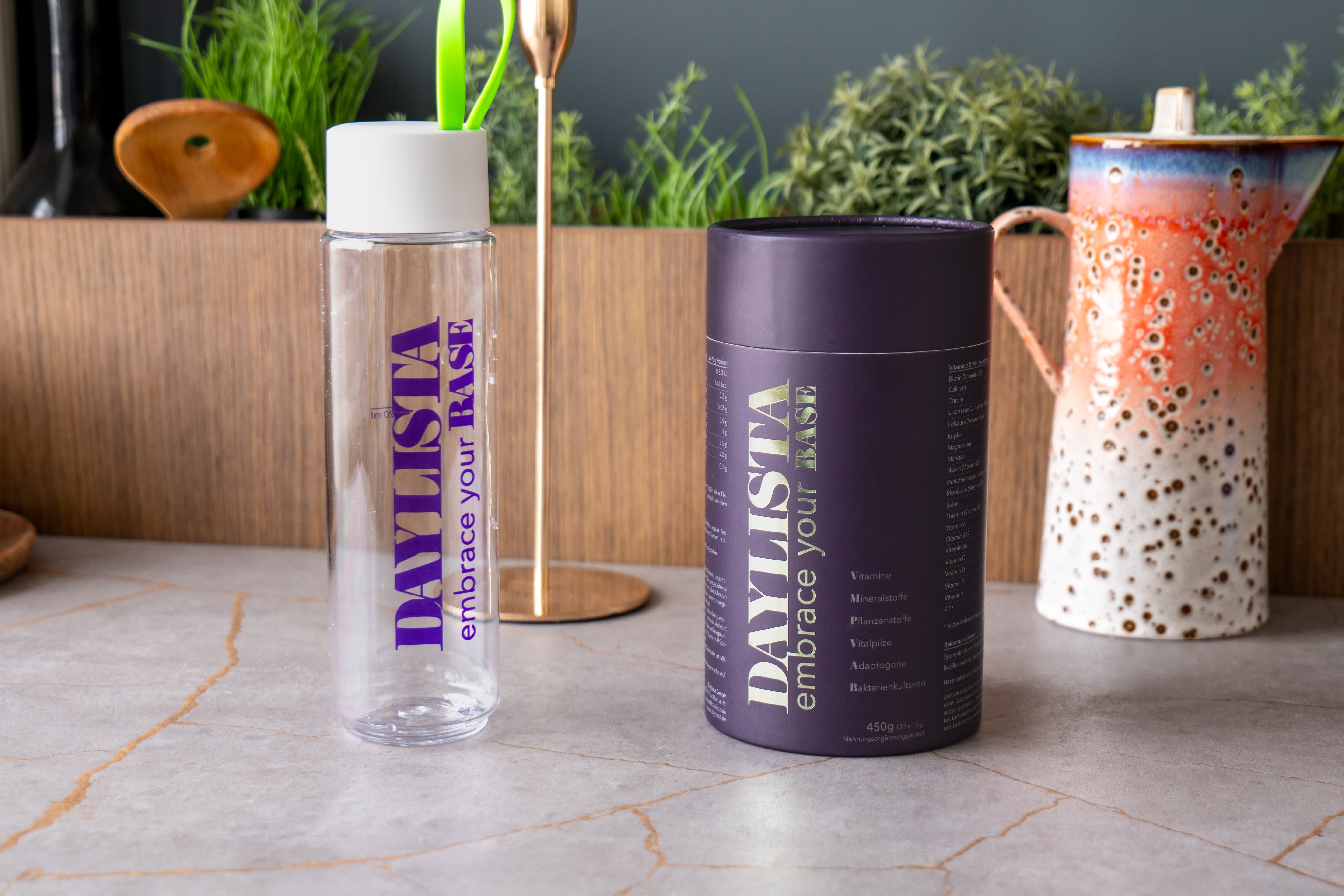 Transparente Daylista-Trinkflasche mit weißem Deckel und grünen Strohhalm steht auf einer Küchenarbeitsplatte neben einer dunkelvioletten Daylista-Dose. Im Hintergrund sind ein bunt gesprenkelter Keramikkrug und Pflanzen zu sehen.