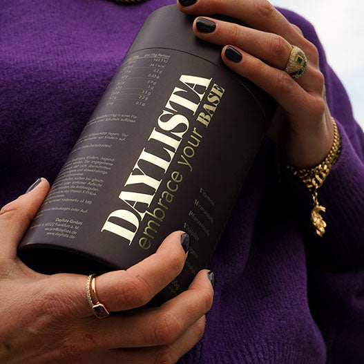 Eine Frau mit goldfarbenem Schmuck und lila Pullover hält eine dunkelviolette Daylista-Dose mit goldenem Schriftzug "embrace your base" in beiden Händen.