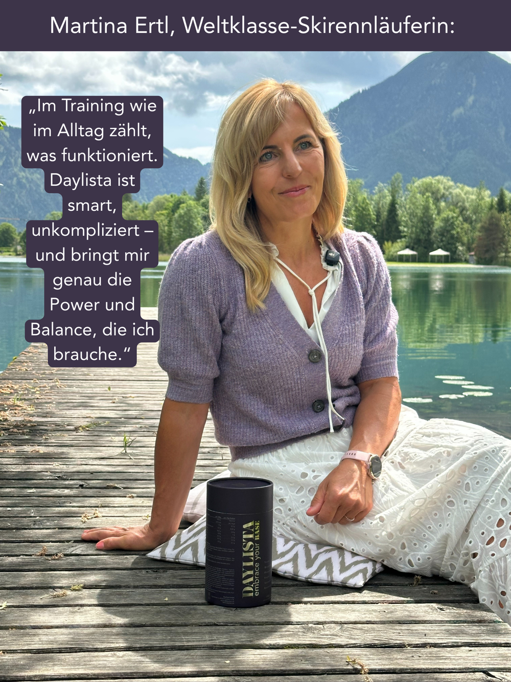 Blonde Frau sitzt entspannt auf einem Holzsteg am See, trägt einen lilafarbenen Cardigan und einen weißen Rock. Vor ihr steht eine dunkelviolette Daylista-Dose; im Hintergrund spiegeln sich Berge und Bäume im Wasser.